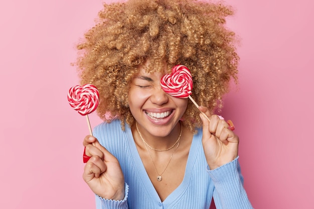 Positive young woman with blonde curly hair hlds two heart shaped caramel candies keeps eyes closed smiles toothily wears casual blue jumper isolated over rosy background Sweet tooth concept