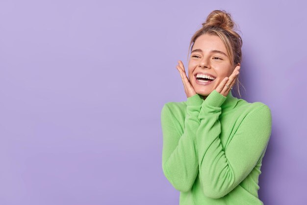 Positive young woman with combed hair keeps hands on cheeks smiles happily feels glad wears casual green turtleneck isolated over purple background copy space away for your advertising content