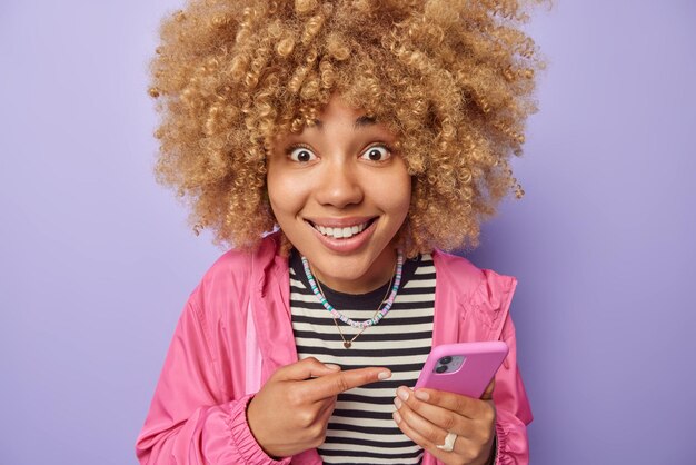 Positive young woman with curly bushy hair smiles gladfully points at her smartphone types sms wears striped jumper and pink jacket isolated over purple background People and technology concept
