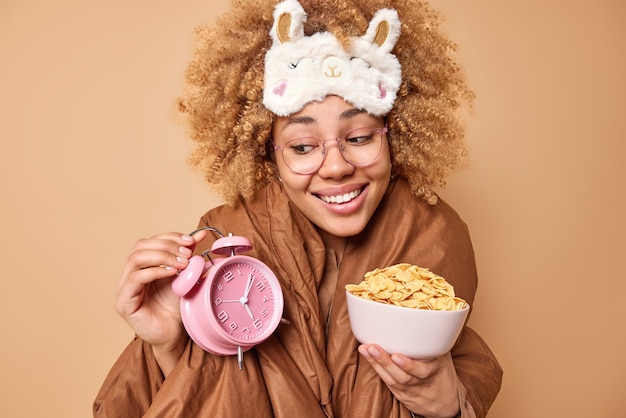 Positive young woman with curly bushy hair wrapped in soft blanket holds alarm clock and bowl of cornflakes being in good mood after awakening isolated over beige background Breakfast concept