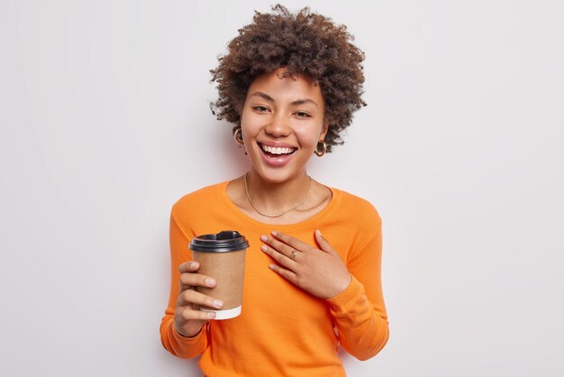Positive young woman with curly hair giggles happily holds disposable cup of coffee enjoys aromatic beverage dressed in casual orange jumper isolated over white background. Drinking concept.