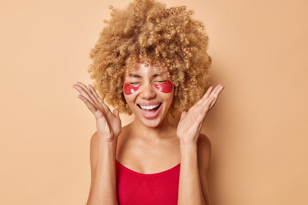 Positive young woman with curly hair keeps palms raised feels overjoyed laughs happily has upbeat mood applies hydrogel patches under eyes for reducing puffiness isolated over beige background