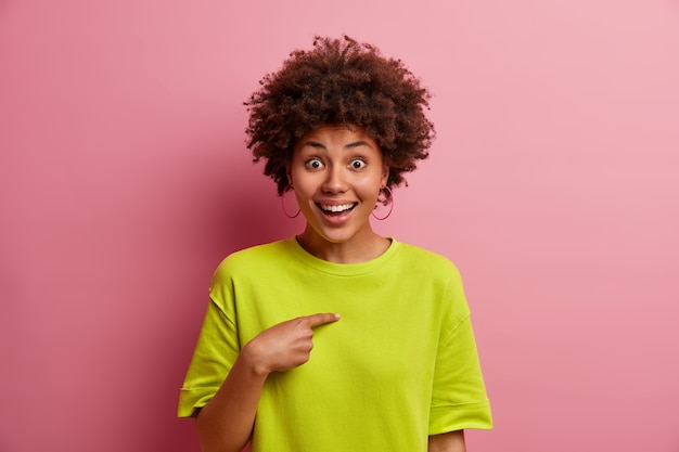 Positive young woman with curly hair points at herself, cannot believe being chosen, picked in someone team, has glad face expression, wears casual green t shirt, isolated over rosy wall