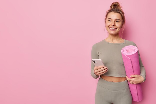 예쁜 머리를 가진 긍정적인 젊은 여성이 롤빵 미소를 지으며 즐겁게 운동복을 입고 분홍색 벽 복사 공간에 격리된 스포츠 뉴스를 읽는 인터넷 서핑에 휴대전화를 사용합니다.