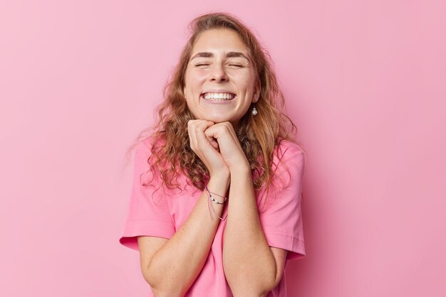 긴 물결 모양의 머리를 한 긍정적인 젊은 여성은 손을 턱 아래에 두고 눈을 감고 카메라를 보며 싱긋 웃으면서 분홍색 배경 위에 격리된 캐주얼한 티셔츠를 입고 즐거운 것을 회상합니다. 행복한 감정 개념