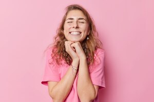 positive young woman with long wavy hair keeps hands under chin closes eyes grins at camera recalls something pleasant wears casual t shirt isolated over pink background. happy emotions concept