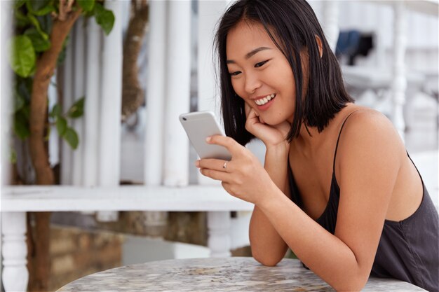 좁은 눈과 단발 머리 헤어 스타일을 가진 긍정적 인 젊은 여성은 소셜 네트워크에서 미디어를 공유하거나 휴대 전화에 새 앱을 설치하고 카페 인테리어에 앉아 있습니다. 온라인에서 긍정적 인 뉴스를 읽는 아시아 여성