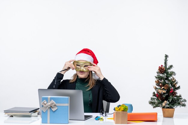 Foto gratuita positivo giovane donna con cappello di babbo natale e indossa una maschera seduto a un tavolo con un albero di natale e un regalo su di esso in ufficio su sfondo bianco