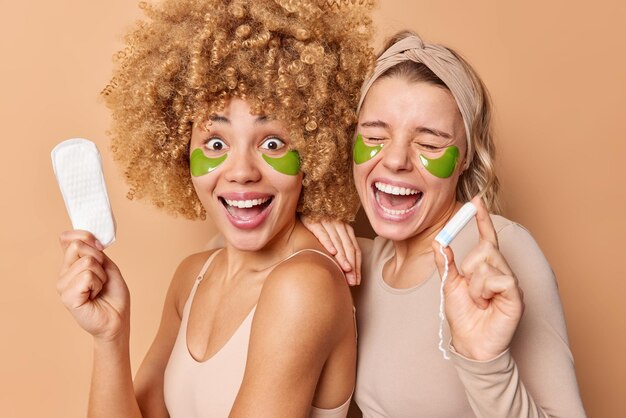 ポジティブな若い女性の笑顔が嬉しそうに立って生理用ナプキンをしっかりと保持し、タンポンが月経に最適な製品を選択しますスキントリートメントのために目の下に緑色のヒドロゲルパッチを適用します美容コンセプト