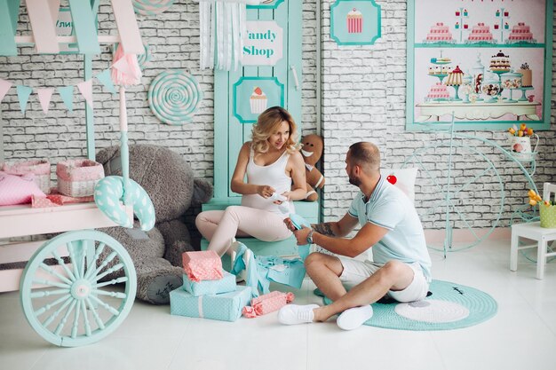 Positivity couple of handsome man and pretty blonde pragnant woman sitting on stairs at studio embracing stomach and gesturing heart by hands Young stylish family at sweet mint interior among toys