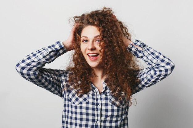 Posittive woman shows her bushy curly hair, wears casual checkered shirt, isolated over white