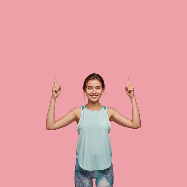 Positve cheerful woman feels glad, points with both index fingers upwards, dressed in casual vest and leggings, models against pink wall. Happy Caucasian girl shows direction upstairs.