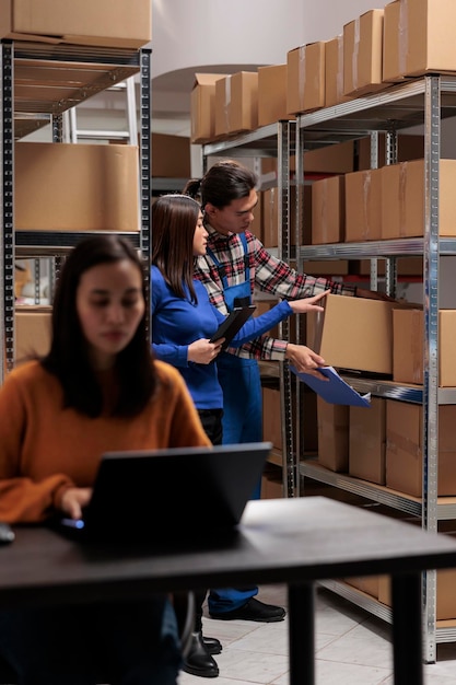 Free photo postal storehouse employees managing order picking and shipping operation postal warehouse asian coworkers taking cardboard box from shelf and analyzing product checklist on digital tablet