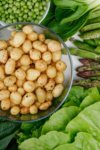 Potatoes in a glass bowl with green pods, peas, spinach, sorrel, lettuce, asparagus top view on a white wall