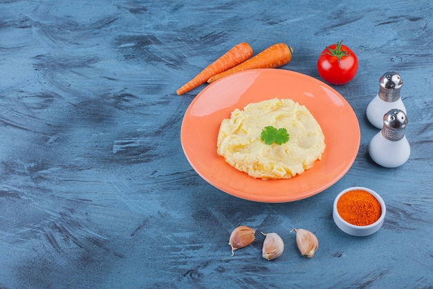 Potatoes puree on a plate next to vegetables and spice bowls , on the blue background. 