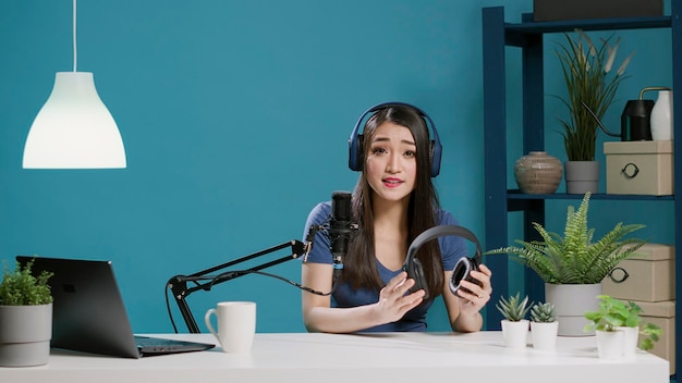 POV of asian woman doing recommendation of modern headphones on vlogging camera, filming product review with wireless headsets. Lifestyle blogger holding earphones. Tripod shot.