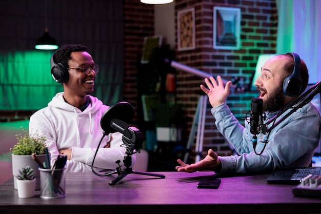 POV of male bloggers vlogging podcast talk on camera, using streaming equipment in studio with neon lights. Content creators having fun filming online episode for social media.