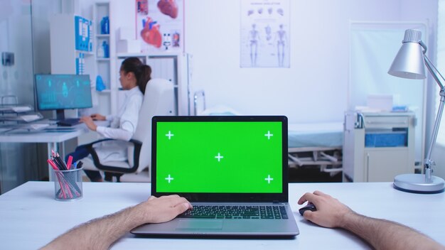 병원 캐비닛에 크로마 키가 있는 노트북을 사용하는 의사와 엑스레이 이미지를 보고 있는 의사의 POV 샷. 의료 클리닉에 녹색 화면이 표시된 노트북을 사용하는 메딕.