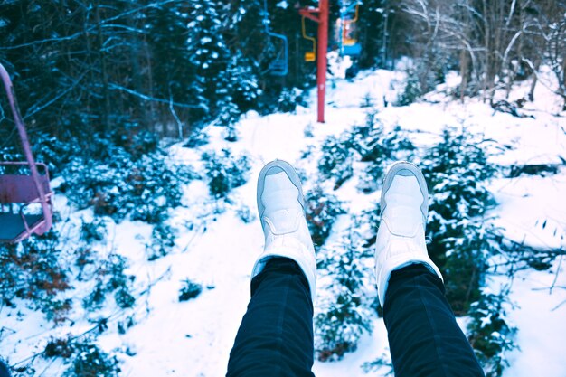POV shot of women legs weared black jeans and white shoes in chair of small grunge ski lift moving through winter forest covered in snow