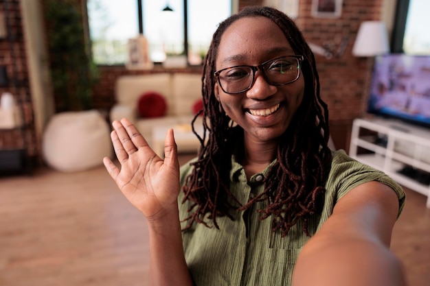 Free photo pov of smiling african american woman waving hello at camera working remote from home living room. friendly student with glasses in video call conference talking with teacher in online class.