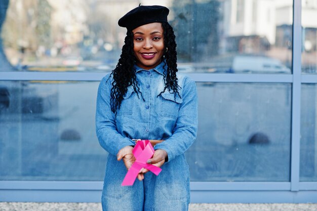 Способность бороться Стильные модные афроамериканки в джинсах и черном берете против современного здания с розовой лентой от рака груди