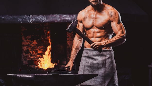 Powerful bearded blacksmith with naked torso and protective apron is standing near working stove with hammer in his hand.