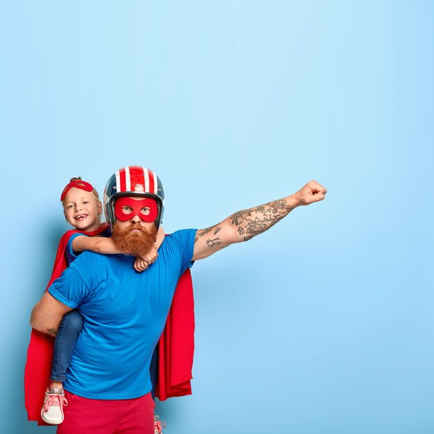 Powerful dad gives piggy back to child, demonstrates courage, makes flying gesture, wears helmet, red mask