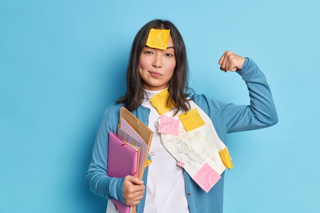 Сильная студентка поднимает руку и показывает мускулы, чувствует себя уверенно после работы над дипломной бумагой, носит наклейки на лбу, держит папки.