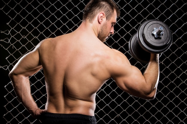 Powerful guy with a dumbbells showing muscles on fence  