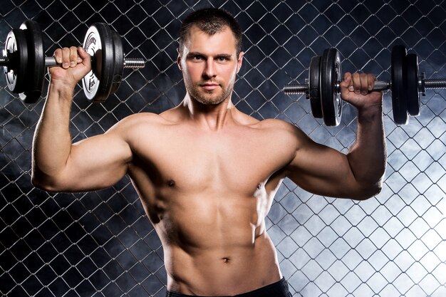 Powerful guy with a dumbbells showing muscles on fence  