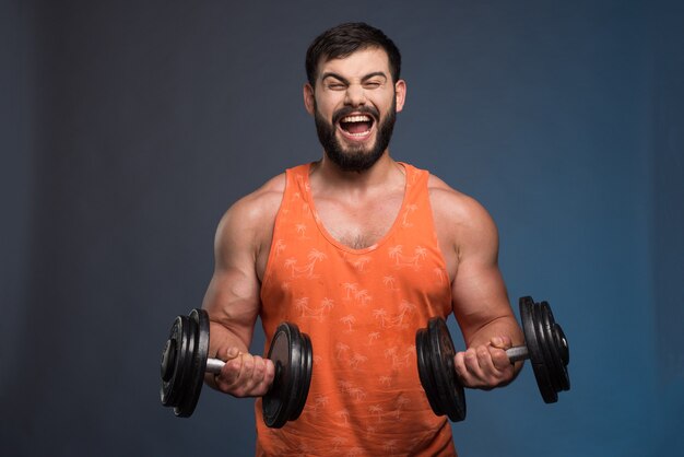 Powerful man holding a dumbbell on dark blue wall.