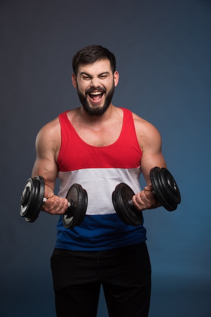 Powerful man holding a dumbbell on dark blue wall.