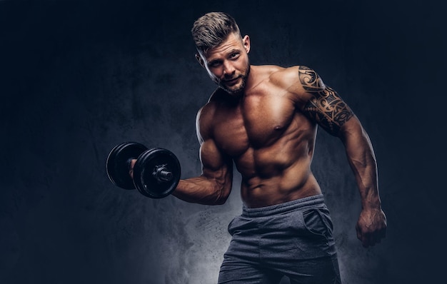 Free photo powerful stylish bodybuilder with tattoo on his arm, doing the exercises on a biceps with dumbbells, look at the camera with a confident look. isolated on a dark background.