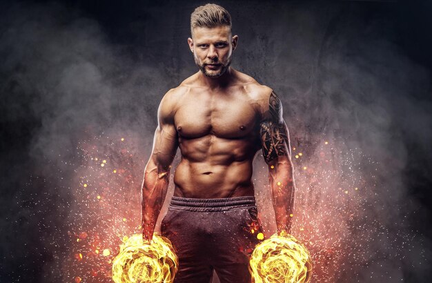 Powerful stylish bodybuilder with tattoo on his arm, posing with dumbbells in a studio, looking at the camera with a confident look. Fire art concept.