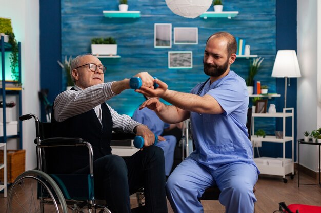 Practitioner man doctor helping retiree senior male in wheelchair to do physiotherapy strength exercise