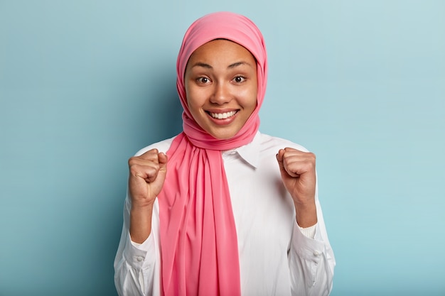 Praying ethnic woman raises clenched fists, rejoices victory, satisfied with good result of work, wears pink hijab, dressed in white shirt, isolated over blue wall. Hand gesture. Happiness.