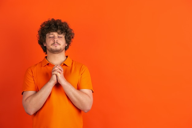 Praying looks calm. Caucasian young man's monochrome portrait on orange  wall. Beautiful male curly model in casual style. Concept of human emotions, facial expression, sales, ad.