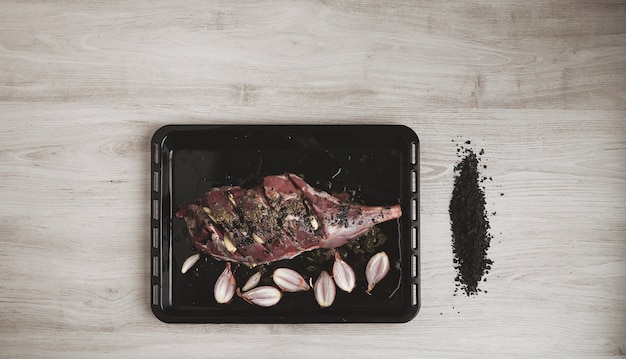 事前に調理されたアイスランドの子羊の脚の肉、スパイス、ハーブ、小さな玉ねぎを黒いベーキングローストディッシュに並べ、火山性の黒い塩を並べて、上面図、白い木製のテーブルで隔離