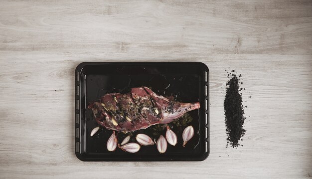 Pre cooked icelandic lamb leg meat wit spices and herbs and small onions on black baking roasting dish and grounded volcanic black salt by side, top view, isolated on white wooden table