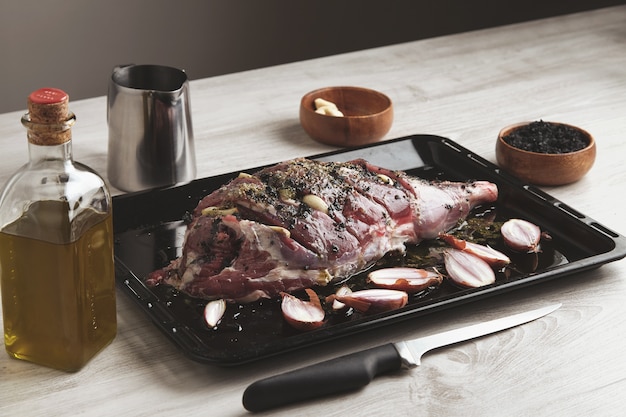 Предварительно приготовленное исландское мясо ягненка со специями и травами и небольшим луком на черной форме для запекания в окружении кухонной посуды, бутылки оливкового масла и деревянной миски с черной солью и ножом впереди