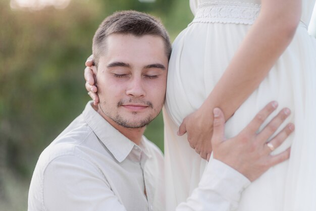 Pregnancy, man is listening to the baby inside woman's belly with closed eyes outdoors