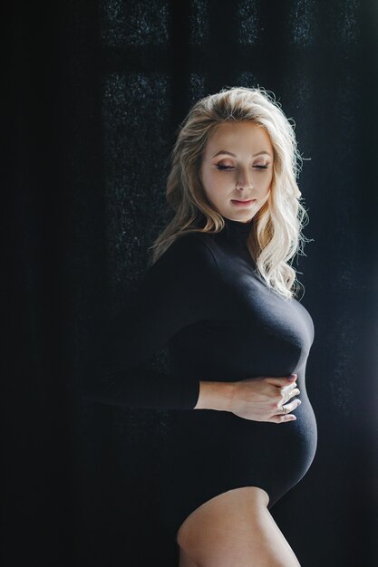 Pregnant blonde woman in a black bodysuit stands near a window and looks down