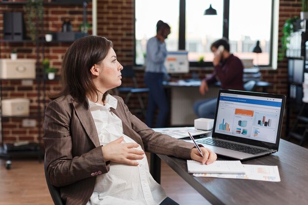 Pregnant businesswoman at office desk using laptop to analyze data charts and project management. Expectant mother reviewing company annual results while sitting in agency workplace.