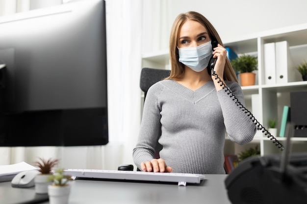 Pregnant businesswoman on the phone at her office desk while wearing medical mask