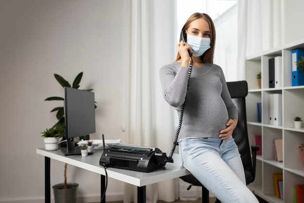 Free photo pregnant businesswoman taking calls in the office while wearing medical mask