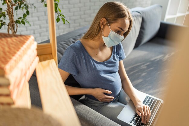 소파에 앉아서 노트북에서 일하는 의료 마스크와 임신 사업가