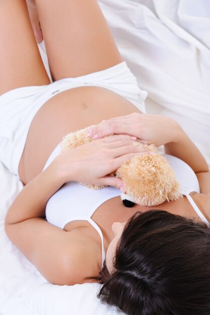 テディおもちゃで白いベッドに横たわっている妊娠中の女性