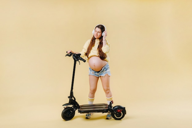 孤立した黄色の背景に電動スクーターの黄色い服とヘッドフォンで妊娠中の女の子。