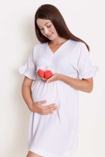 Pregnant happy woman holding red knitted baby shoes in her hands, touching her belly with charming smile