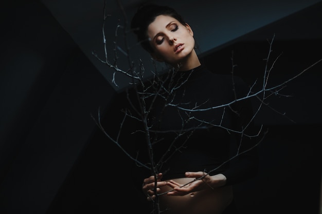 Pregnant woman in black clothes stands under grey wall and holds a tree branch